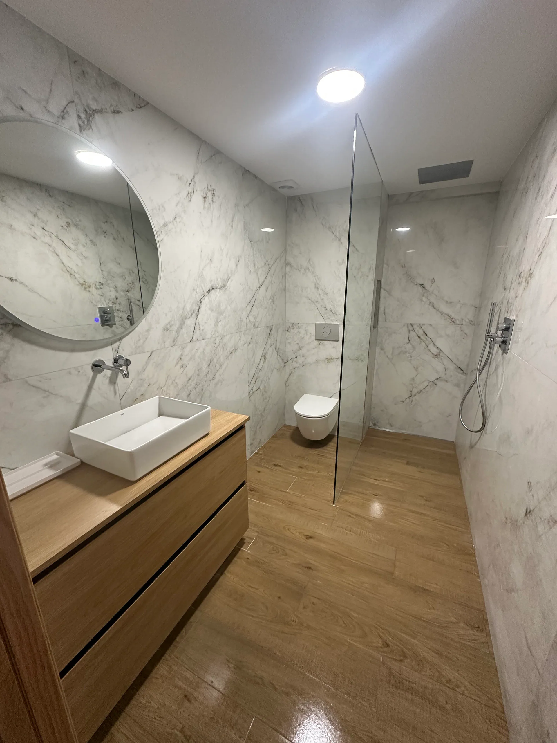 Baño completo revestido en mármol blanco, con lavabo sobre mueble de madera, espejo redondo y ducha de obra con mampara de cristal